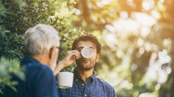A young man drinks coffee out of a mug with an older man